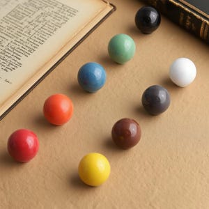 May include: Nine ceramic knobs in a variety of colours, including red, orange, yellow, blue, green, brown, grey, white, and black. The knobs are arranged on a neutral surface, with an open book visible in the background.