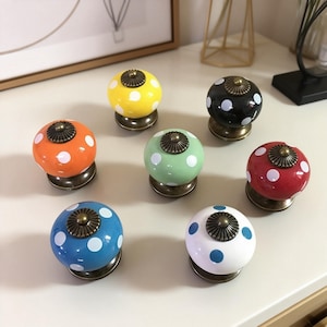 Pode incluir: Uma coleção de sete puxadores de armário de cerâmica coloridos. Cada puxador tem uma cor diferente, incluindo amarelo, laranja, verde, azul, branco, vermelho e preto, e apresenta bolinhas brancas. Os puxadores têm uma base e um detalhe superior em tom bronze.