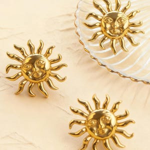 Op de afbeelding: Drie goudkleurige, zonvormige decoratieve objecten. Elke zon heeft een lachend gezicht in het midden met stralende stralen. Eén zon staat op een heldere, geschulpte schaal. De andere twee staan op een lichtbeige oppervlak.