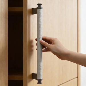 May include: A cabinet door with a light wood finish features a long, textured, gray handle. The handle is mounted on dark metal brackets. The cabinet door is partially open, revealing a dark interior.