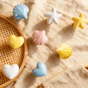 May include: A collection of ceramic decorative knobs in various shapes and colors. The assortment includes blue, pink, and yellow seashells, white and yellow starfish, and yellow, white, and blue hearts. These knobs are arranged on a beige fabric.