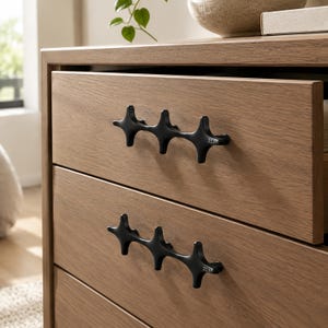 May include: A light brown wooden dresser featuring two drawers, each equipped with a matte black metal handle. The handles have a distinct, interconnected star-like shape with three points and the small engraved text 3219 on the right side.