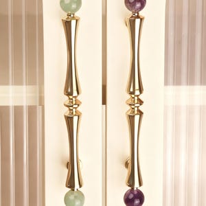 May include: Two cabinet handles with a gold finish. Each handle has a long, ornate design with a gemstone sphere at the top and bottom. One handle has a green sphere, the other a purple sphere.