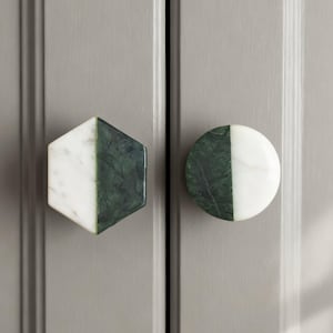 Puede incluir: Dos tiradores de armario, uno hexagonal y otro redondo, cada uno dividido diagonalmente con mármol blanco y verde oscuro. Los tiradores están montados en una puerta de armario gris claro.