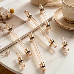 May include: A collection of clear acrylic cabinet pulls with gold-toned metal accents. The pulls are cylindrical with a transparent center and are arranged on a white surface. The hardware is designed for furniture and home decor.