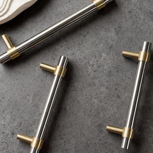 May include: A collection of cabinet hardware featuring a brushed silver finish with gold accents. The T-bar handles have a cylindrical design with a gold ring and a gold T-shaped end. The hardware is designed for easy installation.