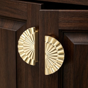 May include: Close-up of two gold-colored, fan-shaped cabinet pulls mounted on a dark brown wooden cabinet door. The pulls have a detailed, textured design and are positioned side-by-side.
