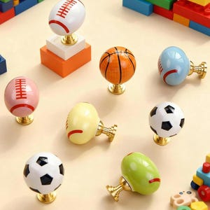 May include: A selection of cabinet knobs with sports ball designs, including footballs, a basketball, and a soccer ball. Each knob has a gold-coloured base. The knobs are set against a neutral background, with colourful toy building blocks visible.