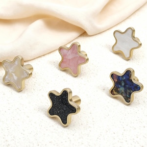Peut inclure: Six boutons de meubles en forme d'étoile avec des bases dorées. Les centres des étoiles présentent diverses couleurs et textures, dont le blanc, le rose, le noir et un motif irisé bleu-vert. Ces boutons décoratifs sont parfaits pour ajouter une touche unique aux meubles.