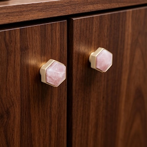 May include: Close-up of two cabinet door knobs. Each knob features a hexagonal rose quartz stone set in a gold-colored metal base. The knobs are mounted on a dark brown wooden cabinet door.