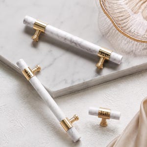 May include: Two long cylindrical cabinet pulls and one small T-shaped knob, each featuring white marble with grey veining and brushed gold metal accents. The gold metal components are engraved with the number 3219 in a clear, dark font.