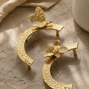 May include: Two gold-colored metal drawer pulls with a hammered texture and a semi-circular shape. One pull features a butterfly ornament, while the other features a flower ornament. The number 3219 is engraved on the end of each handle.