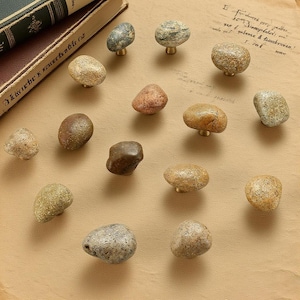 Peut inclure: Ensemble de boutons de meubles décoratifs en pierres naturelles de différentes couleurs, dont le gris, le marron et le beige. Chaque bouton est monté sur une base en laiton. Les boutons sont disposés sur une surface beige.
