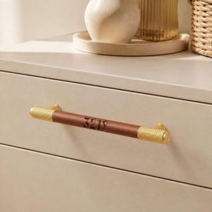 May include: A drawer pull featuring a brown leather center section with the number 3219 embossed in black. The handle has textured gold-toned metal ends and is mounted horizontally on a smooth, light-colored drawer front surface.