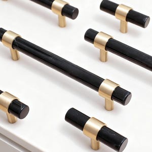May include: Close-up of several black and gold cabinet pulls. The pulls have a cylindrical black handle with a gold-colored ring and base. The hardware is arranged on a white surface, suggesting a display or product presentation.
