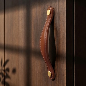 Könnte beinhalten: Ein brauner Leder-Schrankgriff mit weißen Nähten und goldfarbener Hardware. Der gebogene Griff ist an einer dunklen Holzschranktür befestigt. Der Griff ist ein stilvolles Detail für Möbel.