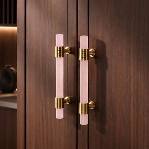 Może przedstawiać: Dwa pionowe uchwyty szafkowe z cylindrycznymi prętami z jasnoróżowego kamienia, mocowane za pomocą polerowanych, złotych metalowych wsporników. Każdy uchwyt posiada dwa okrągłe złote mocowania przytwierdzone do ciemnobrązowych drewnianych drzwi.