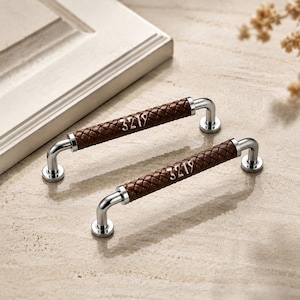 May include: Two cabinet pulls featuring a polished silver metal frame and a brown braided leather grip. Each handle is marked with the number 3219 embossed on the leather surface. The hardware includes two circular mounting bases on each end.