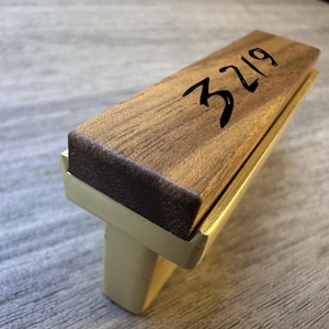 May include: A rectangular drawer pull featuring a dark brown wood top with the number 3219 printed in black ink. The base of the handle is constructed from a smooth, matte-finished light gold metal, providing a contrast to the wood grain.