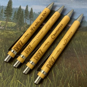 May include: Four wooden pens with silver tips and clips, each engraved with the game title "DAYZ" and various phrases. The pens are arranged diagonally on a green and brown surface, with the game's logo visible in the background.