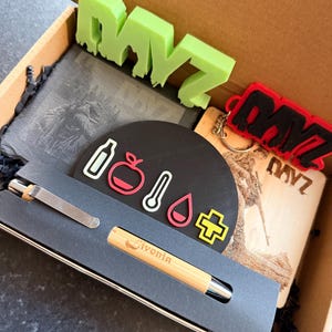 May include: A gift box with assorted items. It features a green "DAYZ" logo, a black coaster with icons, a wooden keychain with a red and black "DAYZ" logo, a slate coaster with an etched image, and a pen with "Livonia" printed on it.