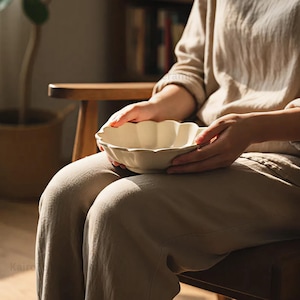 Puede incluir: Un cuenco de cerámica color crema con borde festoneado. El cuenco lo sostiene una persona sentada en una silla de madera. El cuenco tiene un acabado mate y un diseño sencillo. El fondo incluye una silla de madera, una planta y una estantería.