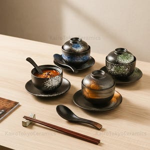 Può includere: Una collezione di ciotole per zuppa in ceramica in stile giapponese con coperchi, piattini e cucchiai. Le ciotole sono nere con accenti bianchi e colorati maculati. Sono presenti anche bacchette e un poggiabacchette. Il testo "KairoTokyoCeramics" è visibile.