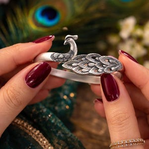 May include: A silver peacock bracelet with intricate feather detailing. The bracelet features a detailed peacock design with a curved body and a raised head. The background includes peacock feathers and a dark green fabric.