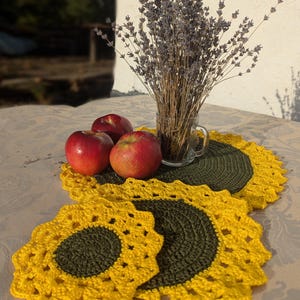 Puede incluir: Naturaleza muerta con tres manzanas rojas, un vaso de lavanda seca y dos manteles individuales de girasol de ganchillo. Los manteles individuales tienen un diseño de pétalos amarillos con un centro verde oscuro.