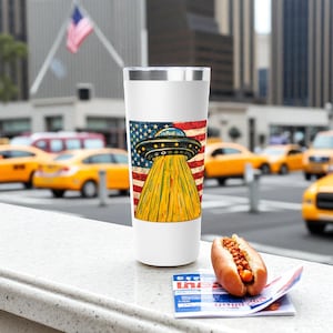 以下が含まれることがあります： 銀色の蓋が付いた白いトラベルマグには、アメリカ国旗を背景に黄色い光を放つUFOのグラフィックが描かれています。 ホットドッグと新聞が前景にあります。