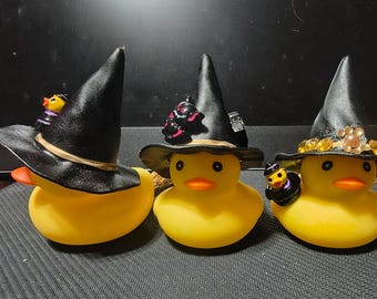 May include: Three yellow rubber ducks wearing black pointed witch hats. The left duck has a tiny duck on its hat. The center duck features a black cat decoration on its hat. The right duck has a floral embellishment and a tiny duck on its hat.