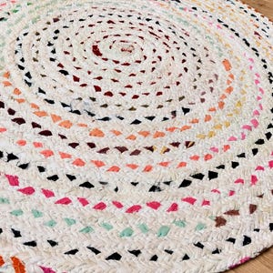 Puede incluir: Una alfombra redonda trenzada con un patrón en espiral construido a partir de tiras de tela blanca intercaladas con acentos coloridos en tonos rosa, naranja, amarillo, azul claro y negro, creando una superficie texturizada con un tejido uniforme.
