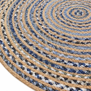 Può includere: Un tappeto rotondo intrecciato caratterizzato da cerchi concentrici di fibre di juta color marrone chiaro naturale alternate a strisce di tessuto denim blu in varie tonalità di indaco chiaro, medio e scuro, creando un motivo circolare testurizzato.