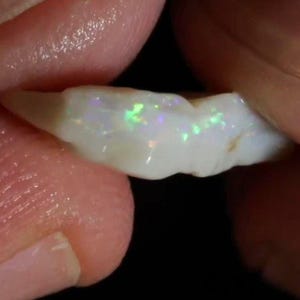 May include: A raw, elongated opal gemstone with a milky white base color and iridescent flashes of green, blue, and pink light visible across its uneven, polished surface. The stone has a natural, irregular shape with a smooth, glossy texture.