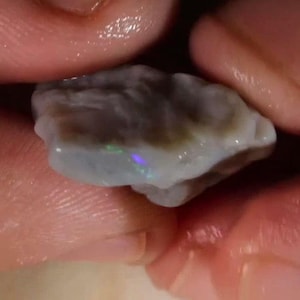 May include: A raw, unpolished opal gemstone with a translucent, milky white base and irregular, undulating edges. The surface displays a subtle, shimmering play of color with small, vibrant flashes of green and purple light visible within the stone.