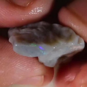 May include: A raw, unpolished opal gemstone with a translucent, milky white base color. The stone features irregular, undulating surfaces and displays subtle flashes of iridescent green and blue light across its textured, organic form.