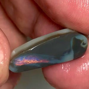 May include: A polished opal gemstone with a dark blue base color and iridescent flashes of orange, purple, and blue. The opal has a smooth, curved shape and a light-colored edge. The gemstone is held between two fingers.