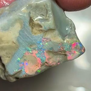 Könnte beinhalten: Ein roher Opal-Edelstein mit hellblauer Grundfarbe und lebendigen roten, rosa, grünen und blauen Blitzen. Der Stein hat eine natürliche, unebene Textur und eine polierte, glänzende Oberfläche. Ein einzigartiges Naturjuwel.