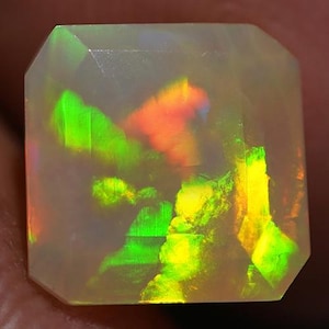 Puede incluir: Una piedra preciosa de ópalo tallada en forma cuadrada con un cuerpo translúcido. El ópalo muestra colores internos vibrantes, incluyendo verde, amarillo y naranja, creando un efecto de juego de colores. Las facetas de la gema capturan y reflejan la luz.