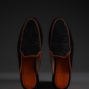May include: A pair of black suede slip-on shoes with a crocodile-textured leather vamp. The shoes have an orange leather lining and trim. The shoes are displayed on a gray surface against a dark background.