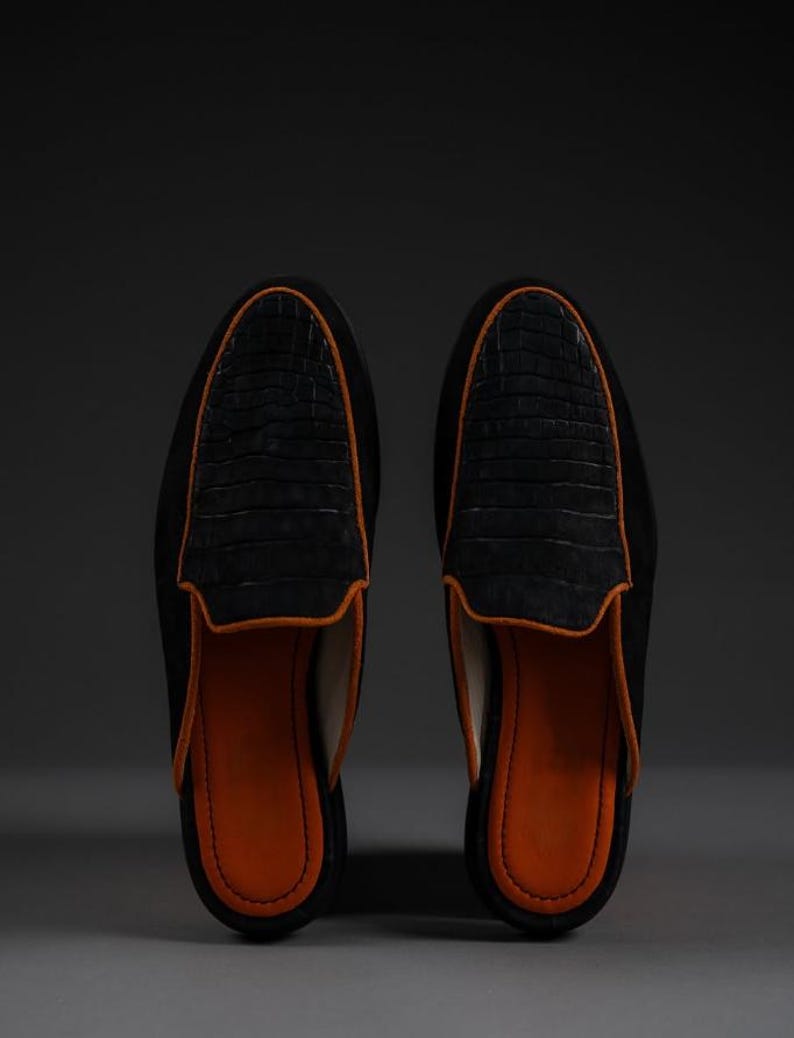 May include: A pair of black suede slip-on shoes with a crocodile-textured leather vamp. The shoes have an orange leather lining and trim. The shoes are displayed on a gray surface against a dark background.