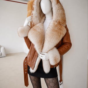 May include: A tan leather jacket featuring a large, plush, cream and light brown fur-lined hood, collar, and cuffs. The jacket includes a matching leather belt with a silver buckle at the waist and long sleeves with zippered details on the sides.