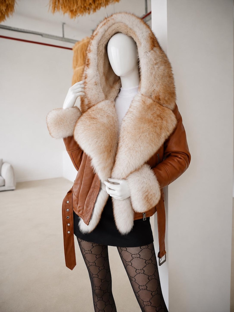 May include: A tan leather jacket featuring a large, plush, cream and light brown fur-lined hood, collar, and cuffs. The jacket includes a matching leather belt with a silver buckle at the waist and long sleeves with zippered details on the sides.