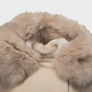 Puede incluir: Primer plano de una chaqueta beige con un gran cuello de piel suave de color marr&oacute;n claro. La chaqueta tiene una cremallera y una textura suave similar al cuero. El cuello de piel a&ntilde;ade un elemento lujoso y c&aacute;lido.