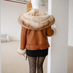May include: A tan leather jacket featuring a hood lined with cream-colored faux fur and a thick brown faux fur trim. The jacket includes matching faux fur cuffs at the sleeves and a belted waist detail, worn over a black skirt and patterned tights.
