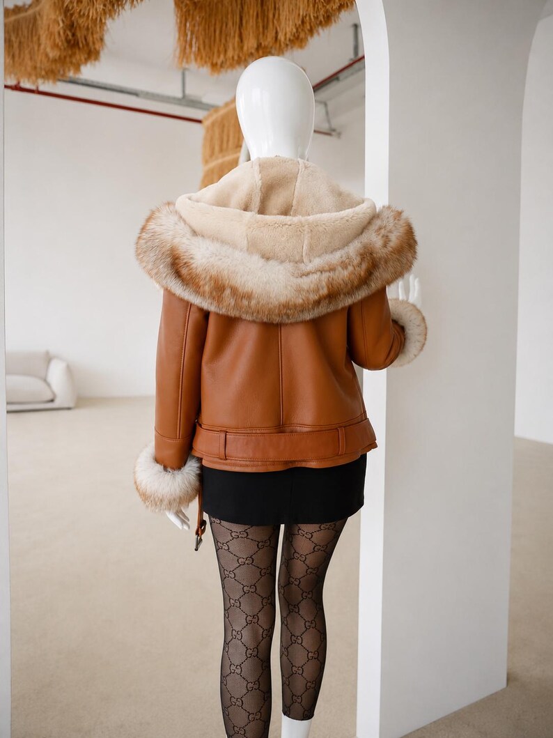 May include: A tan leather jacket featuring a hood lined with cream-colored faux fur and a thick brown faux fur trim. The jacket includes matching faux fur cuffs at the sleeves and a belted waist detail, worn over a black skirt and patterned tights.