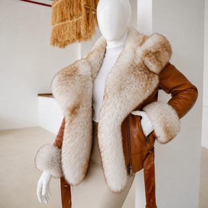 May include: A tan leather jacket featuring a large, thick cream and light brown fur collar and matching fur cuffs. The jacket includes a front zipper closure, a belted waist with a metal buckle, and long sleeves, displayed on a white mannequin.