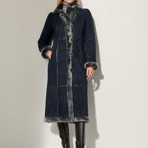 May include: A long, navy blue coat with gray fur trim and lining. The coat features a button-up front, a wide collar, and cuffs trimmed with fur. The model is wearing black knee-high boots. The coat is a stylish outerwear option.