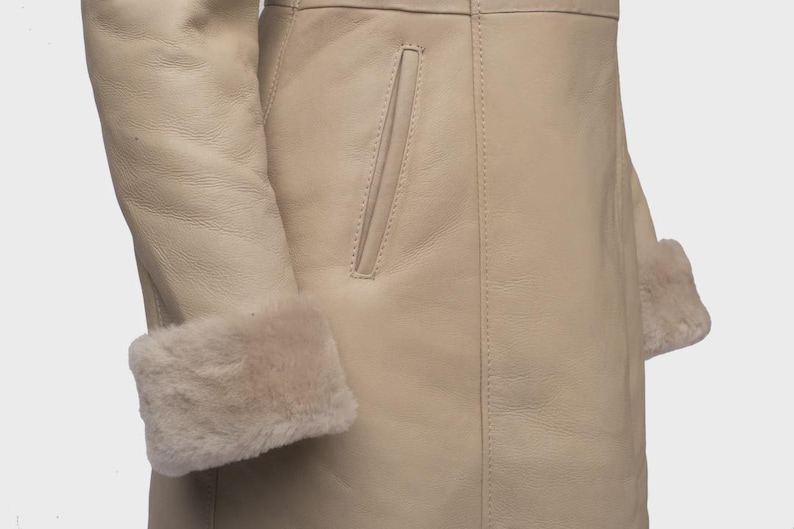Puede incluir: Primer plano de un abrigo de cuero color crema con ribete de borrego en los pu&ntilde;os. El abrigo presenta un bolsillo y detalles de costura visibles. El dise&ntilde;o es sencillo y elegante.
