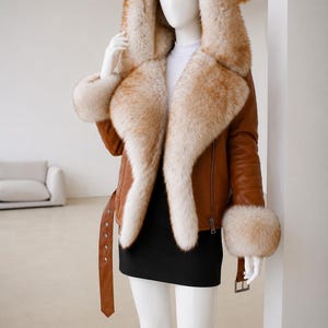 May include: A tan leather jacket featuring a large hood, wide lapels, and cuffs trimmed with thick, fluffy, light brown and cream-colored fur. The jacket includes a matching leather belt with a silver-toned buckle and a side zipper closure.